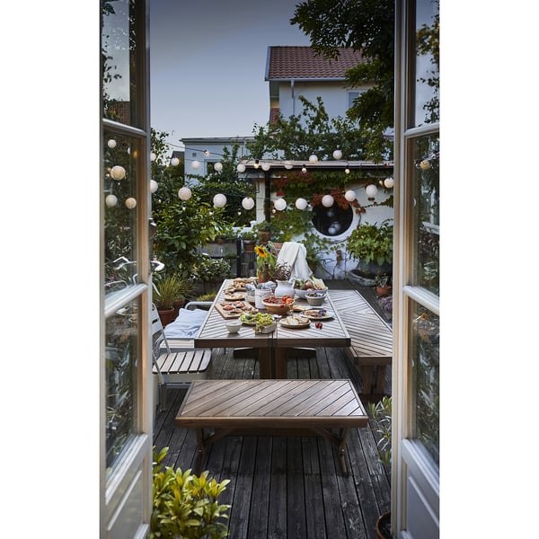 Outdoor wooden dining area with NORRMANSÖ bench. Round tables with food and hanging lights.