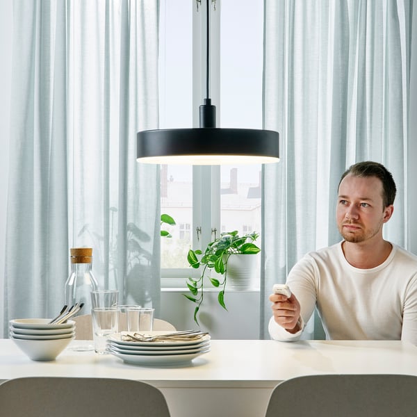 A person holds a remote, controlling a modern, hanging lamp above a dining table with dishes and glasses.