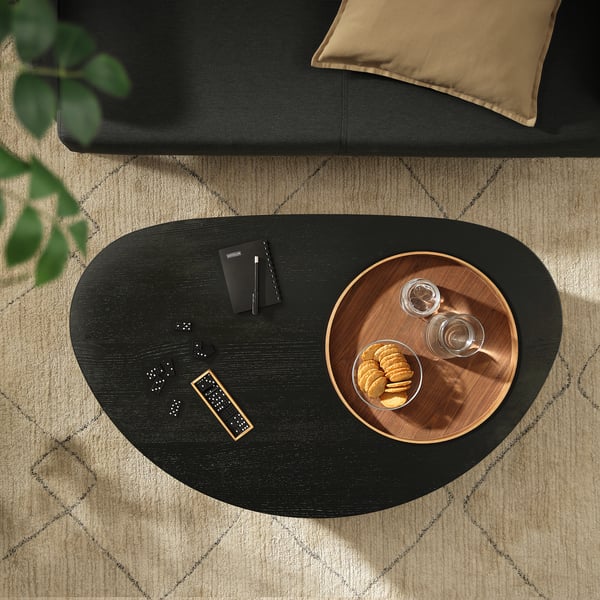 A black ash ÖRTRÄSK oval coffee table with remote, snacks, and notebook on a rug.