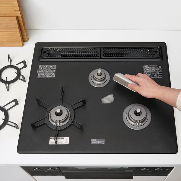 Hand cleans black stove with three burners, one with fish grill, set on white counter.