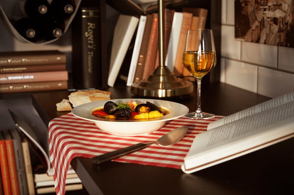 Cosy dining setup: white bowl of colourful mussels next to wine glass on red striped cloth, surrounded by books.