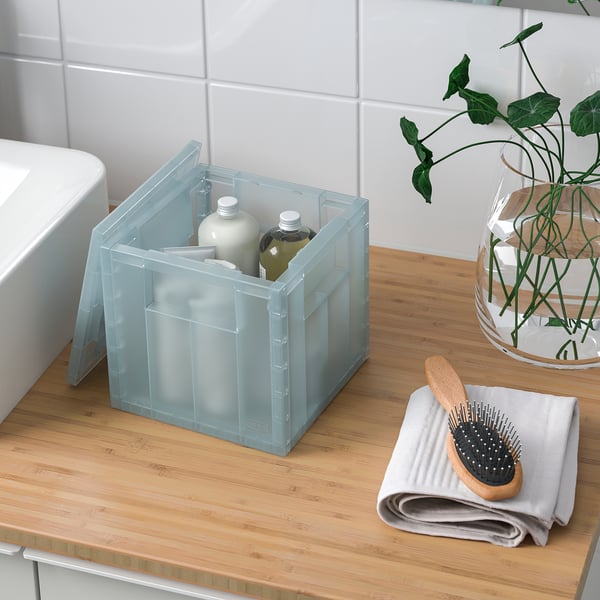 Clear plastic storage box with lid on wooden counter, holding shampoo and conditioner bottles, next to folded towel and hairbrush.