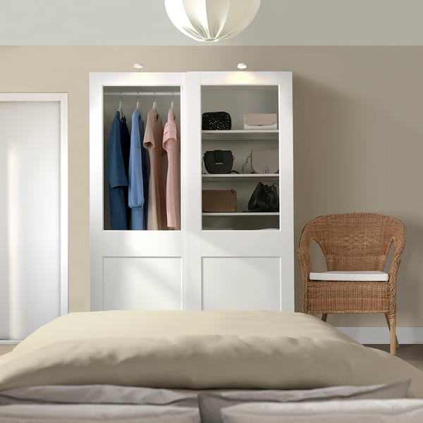 The PAX wardrobe in white, housing neatly arranged clothes with glass panels. It stands tall beside a wicker chair and a white bed.