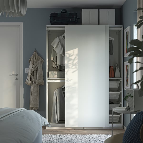 A modern bedroom with a white PAX wardrobe featuring sliding doors, a hanging jacket, shelves, and a drawer with a brown bag.