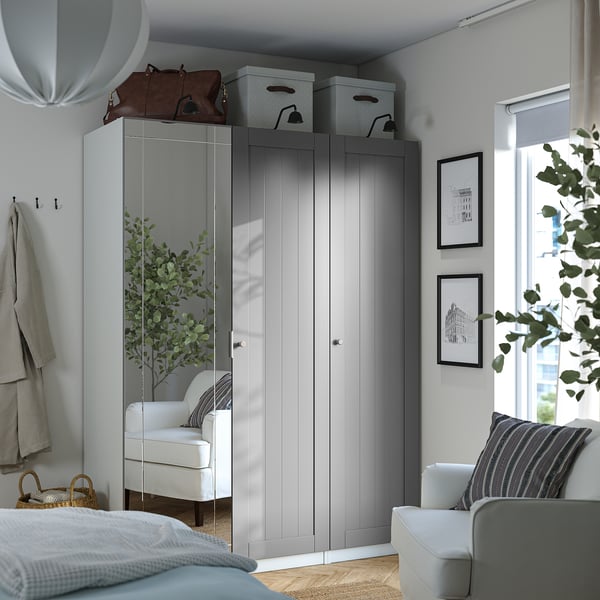 Modern bedroom with large grey PAX wardrobe, tall white chair, bed, and wall decor. Wardrobe is big, mirrored, with boxes on top.