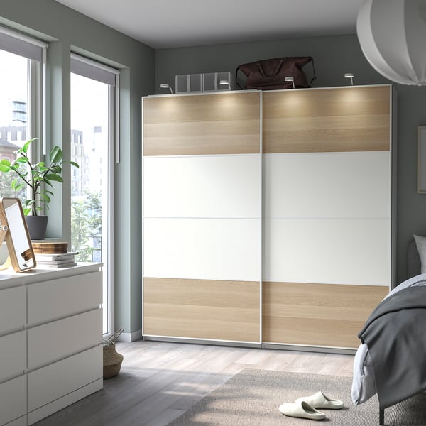Modern bedroom with white and light wood PAX wardrobe, sliding doors, grey walls, large window, plant, and bed.
