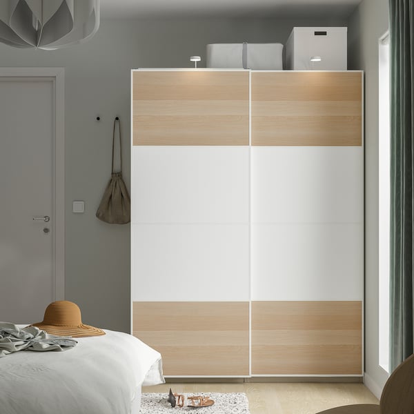 Modern PAX wardrobe with white sliding doors and light wood next to bed. Top shelf, hat on wall, cosy bed with woven hat.