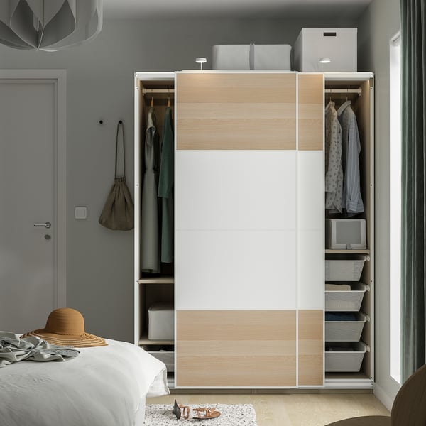 Modern bedroom with open PAX wardrobe displaying clothes, baskets, and storage boxes. Sliding doors and adjustable feet are visible.