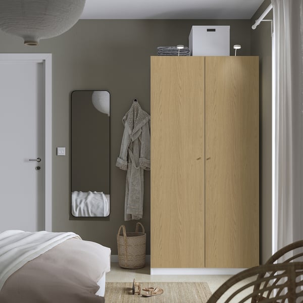 Minimalist bedroom with tall wood PAX wardrobe, wicker basket, grey robe, black mirror, white door, and bed with white linens.