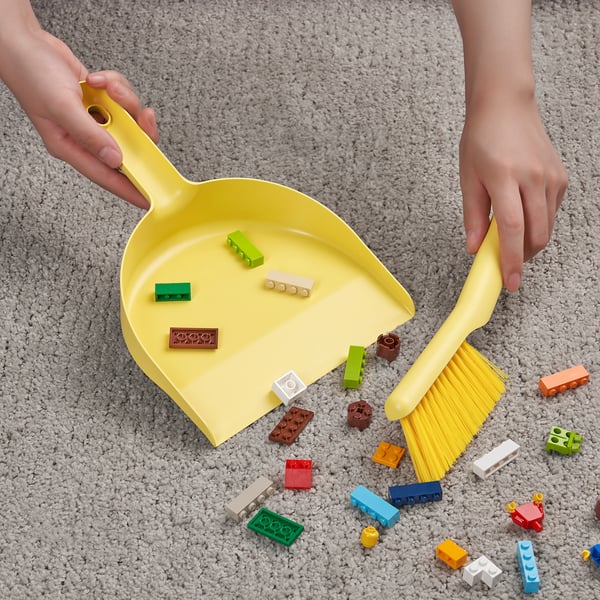 A child holds a yellow dustpan and brush, collecting colourful toy blocks scattered on a carpeted floor.