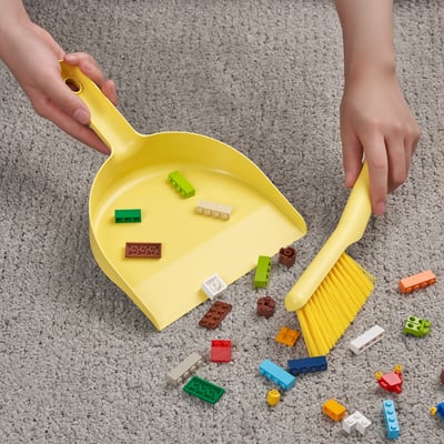 A child holds a yellow dustpan and brush, collecting colourful toy blocks scattered on a carpeted floor.