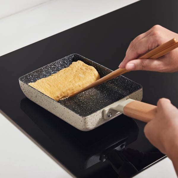A hand uses chopsticks to roll an omelette in a small, grey non-stick frying pan on a black stovetop.