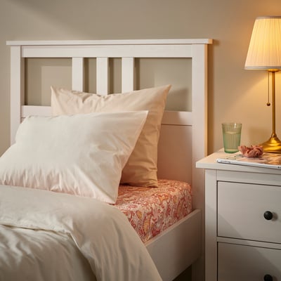 White bed with peach pillows, paisley-patterned fitted sheet. Brass lamp & green glass on bedside table.