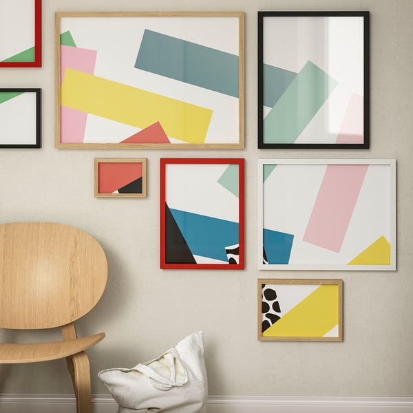 Five colourful abstract art frames in geometric shapes and pastel colours, varied sizes and borders, displayed on a wall with a wooden chair.