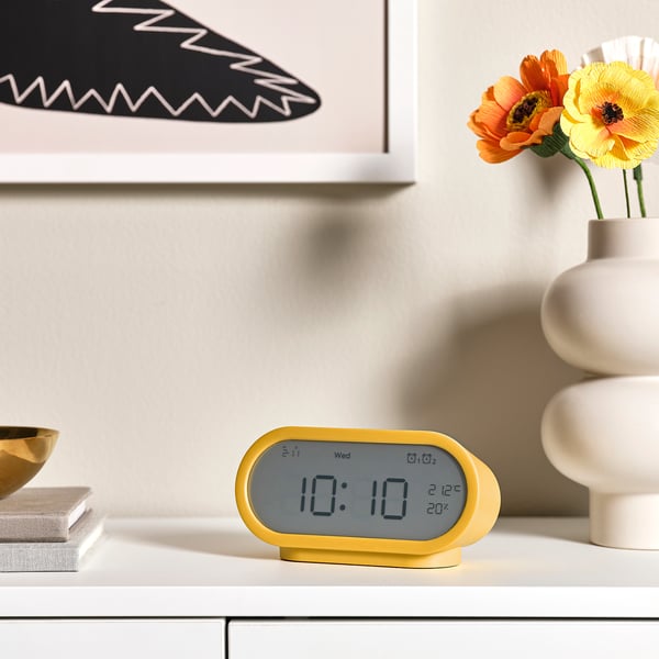 Yellow RÖDSTRÄFSE alarm clock with time, alarm, temp, humidity on table. Orange flowers, abstract art behind.