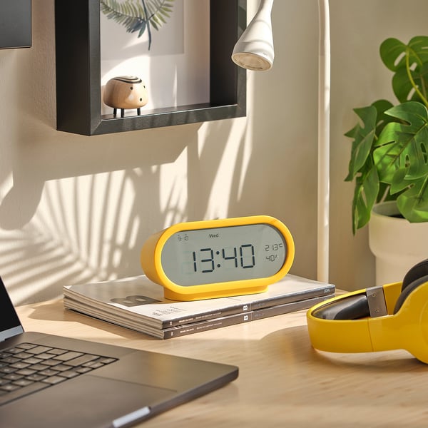 Yellow digital alarm clock on desk with laptop, cds, headphones, plant, and wooden figure.
