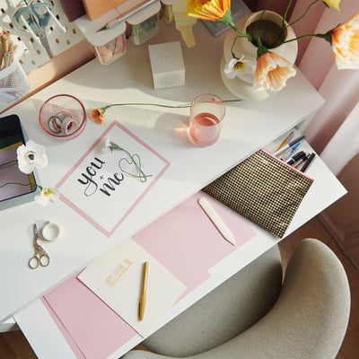 A stylish workspace with a white desk featuring a you + me card, pink accessories, flowers, and a small beige ROMELSÖN bag near notepads and pens.
