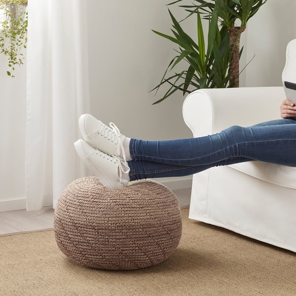 Person sitting on white chair, feet up on round knitted pouffe. Light-filled room with plant.