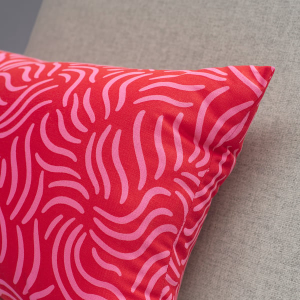 A vibrant red cushion with pink swirls rests on a soft grey surface, showcasing its high-quality fabric and plush design.