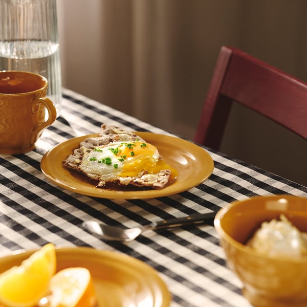 Yellow plate with fried egg on toast, surrounded by cups and bowls on a black-and-white chequered tablecloth.