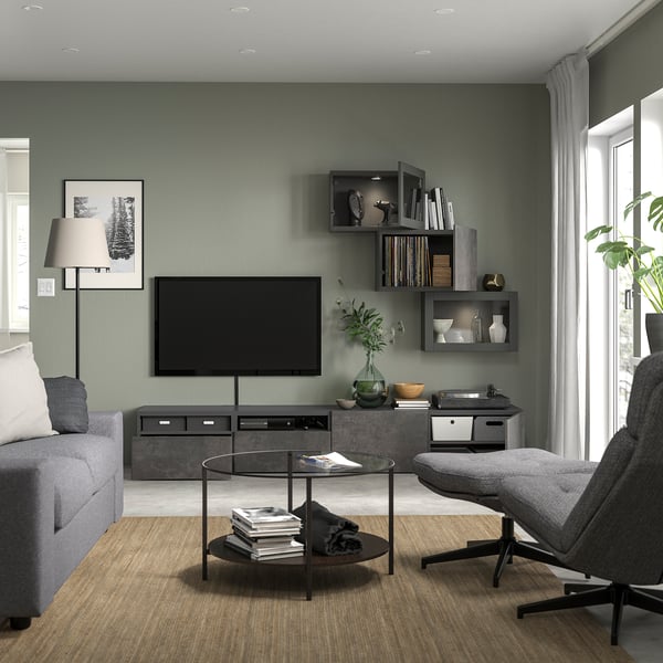 Modern living room with grey walls, grey sofa, large tv, glass round coffee table with books, grey armchair, plant, and built-in shelves.