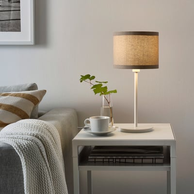 Modern living space with SKAFTET white lamp on small table, next to cosy sofa with blanket and decorative cup.