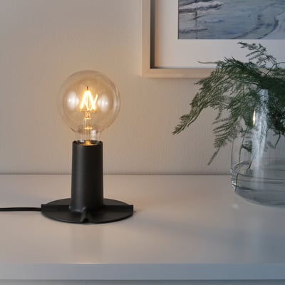 Table lamp SKALLRAN with warm bulb casting light on surface. Glass vase with greenery beside it against wall.