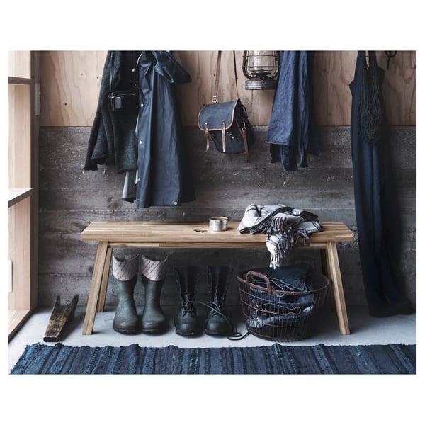 Simple entryway with light wood bench, black boots, coats, and blue bag. Wall hooks and rug.