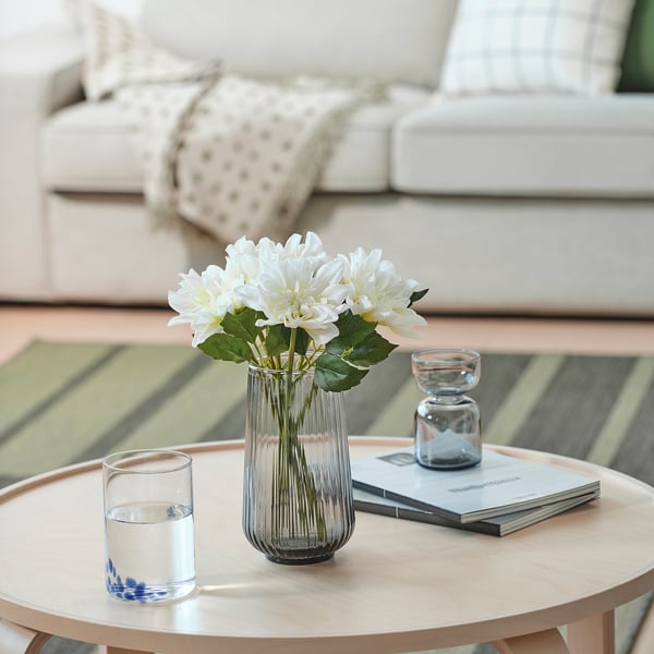 White artificial dahlia in clear vase on wooden table with books, glassware, and candle holder. Background: white sofa with pillow.
