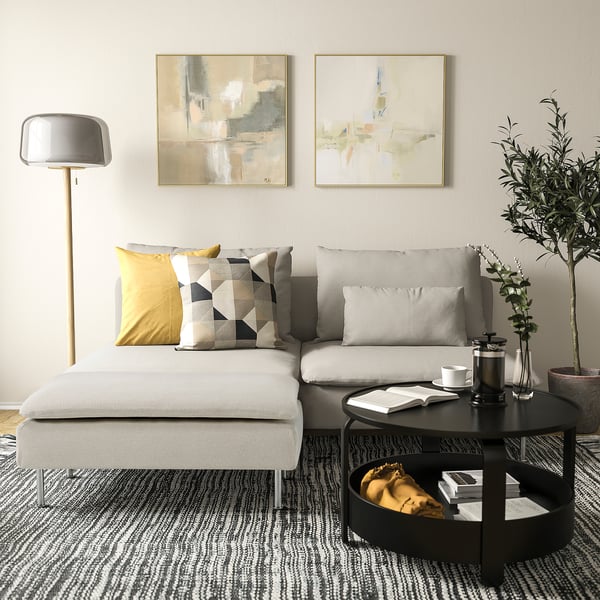 Modern living room with white SÖDERHAMN sofa and footstool, grey pillows, black coffee table, lamp, and abstract paintings on wall.
