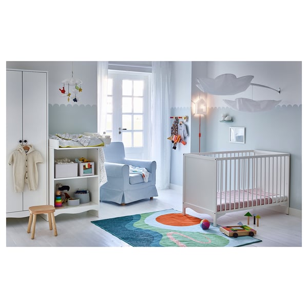 Baby nursery with white cot, changing table, blue armchair, toy shelf, hanging mobile, rug, and wood stool.
