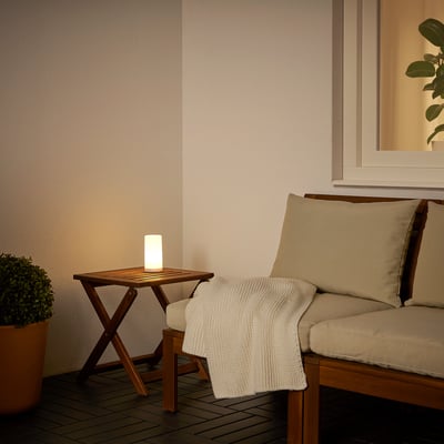 Small white outdoor lamp on wooden table near cushioned bench, plant, and wall.