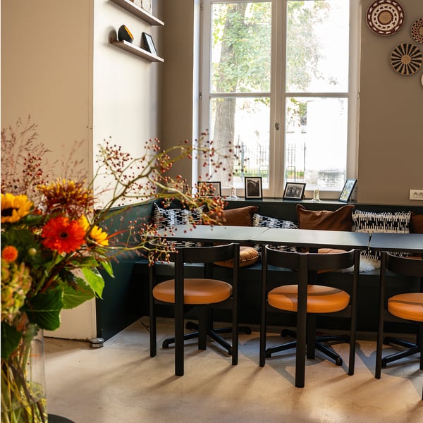 Bright café with STENSELE table in green, square with 4 black stools, positioned near large windows, colourful flowers in foreground.