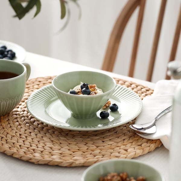 Small green bowl filled with ice cream, blueberries, and nuts sits on a matching green plate on a woven placemat, part of the STRIMMIG series.