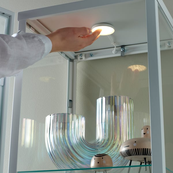 A hand presses a embedded cabinet light, showing its on/off function. The light illuminates a mirrored cabinet containing decorative items.