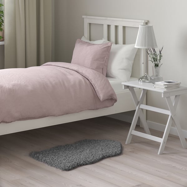 Bedroom scene with white bed, pink linens, white bedside table, lamp, plant, and dark grey SVINDINGE rug, smaller than the bed.