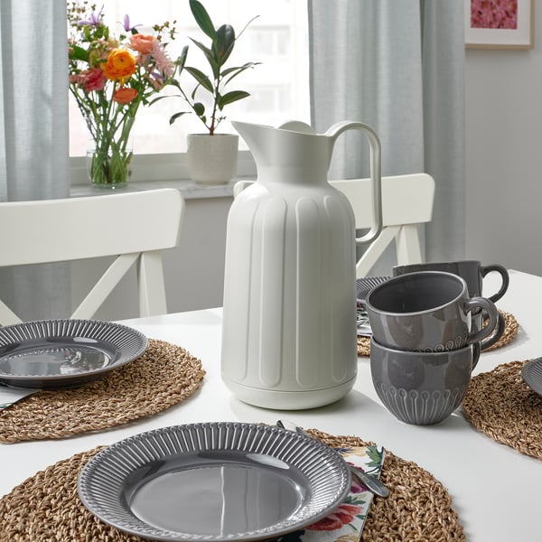Table set with grey plates and cups, white vacuum jug, and woven placemats. Flowers and plant on windowsill.