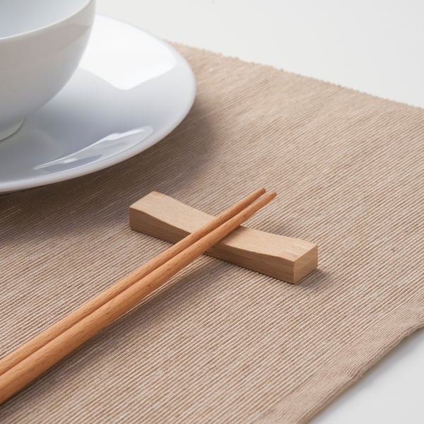Chopsticks resting on wooden holder, beside a white cup on beige placemat.