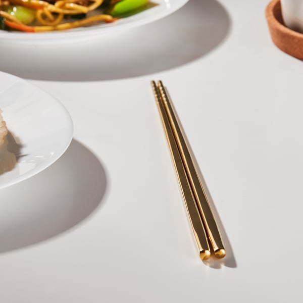 Gold chopsticks on a table with dishes. Easy to grip, non-slip, and square-shaped. Part of the TILLAGD series.