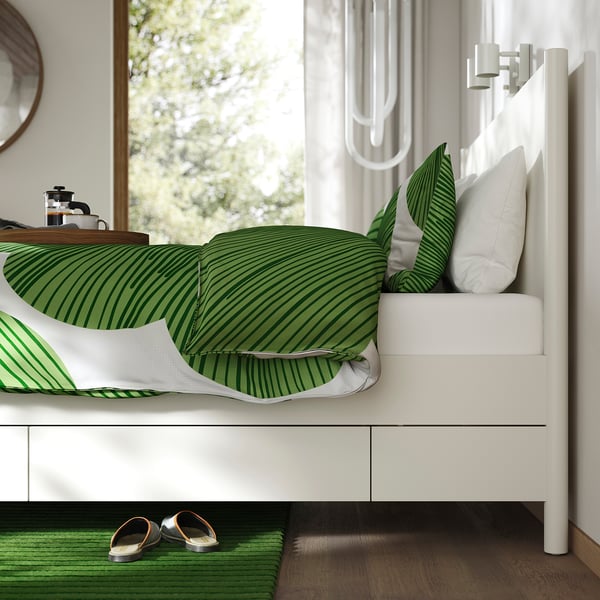 A white TONSTAD bed frame with soft-closing drawers, showcasing green striped bedding and pillow, with slippers placed on a green rug beside it.