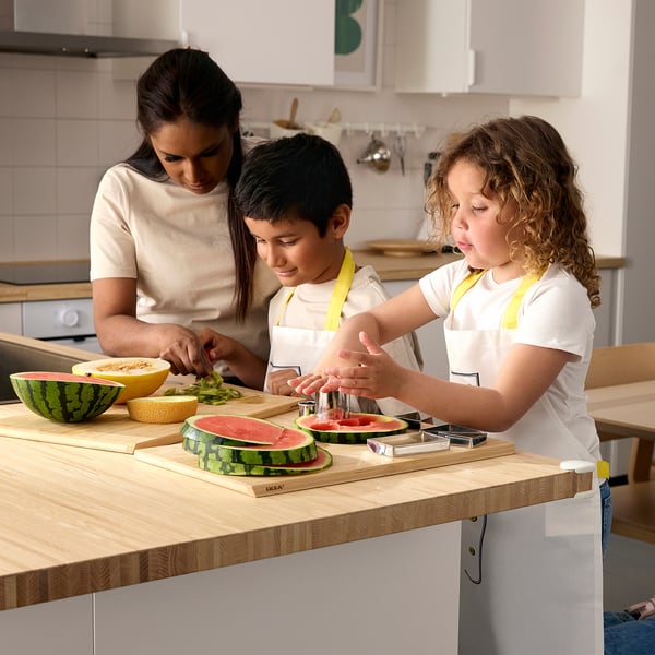 Children and mum prepare food. Kids wear aprons and hats, safely cutting watermelon.
