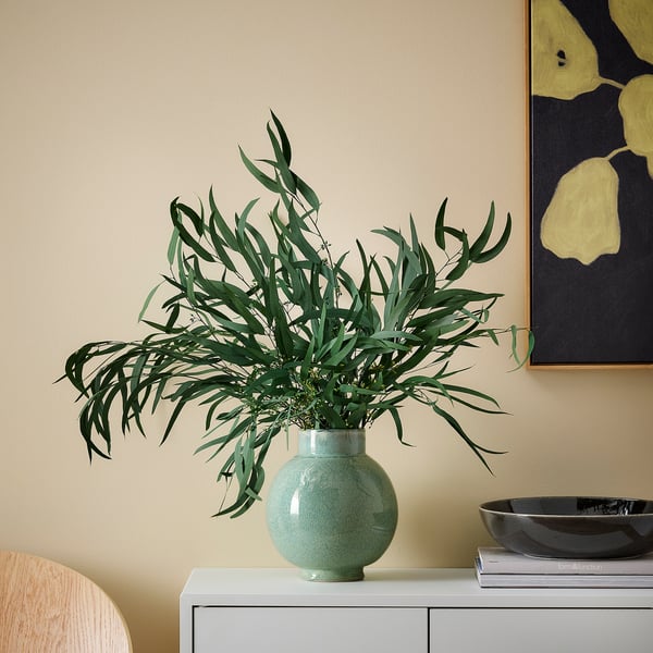TORKA green vase with dried plants on white sideboard, black bowl, books, chair, yellow-black wall art.