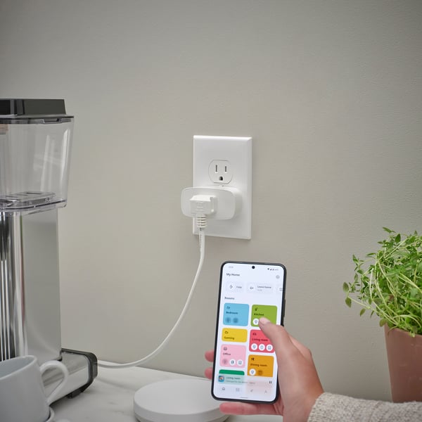 Hand holds smartphone controlling smart plug via app, plugged into kitchen wall outlet.