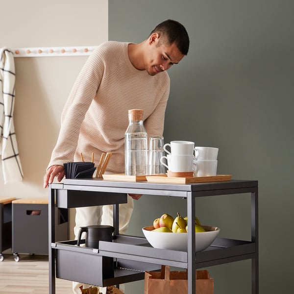 Person in beige sweater organises items on a black TROTTEN trolley, which holds mugs, water bottles, and a bowl of apples.