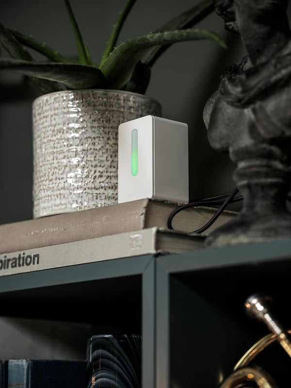 A small white sensor with a green light sits on books, next to a potted plant.