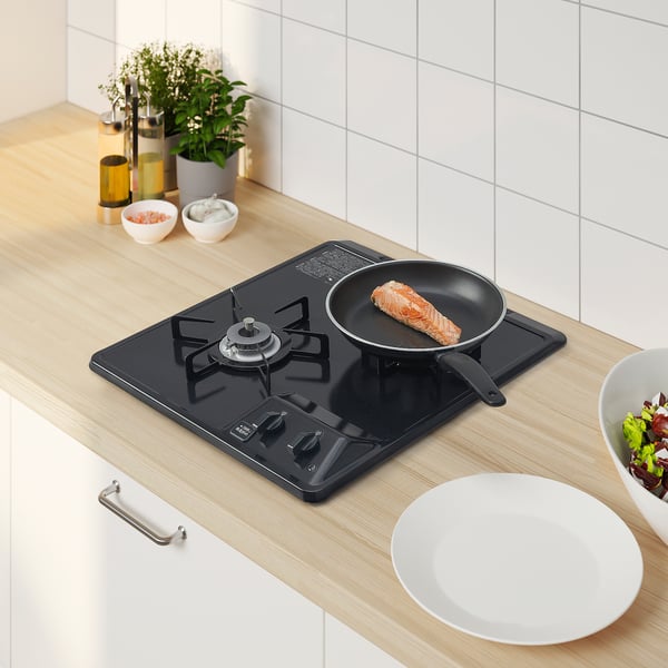 Small, black gas hob, VISSNA, with two burners cooking fish. White dish, bowls, and plant nearby on light countertop.