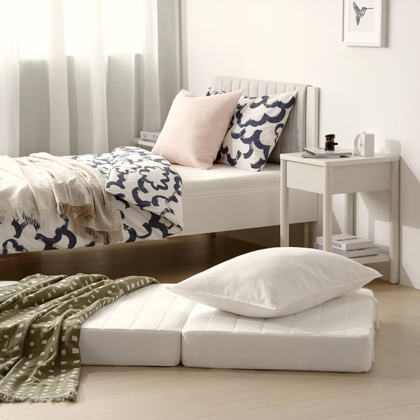 Modern bedroom with white bed frame, mattress, pillows, blankets, and side table. Light-colored wooden floor with minimalist decor.