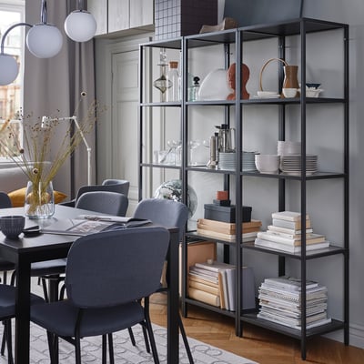 Modern dining room with metal & glass bookshelf, plates, vases, books. Grey chairs around table with floral centrepiece.
