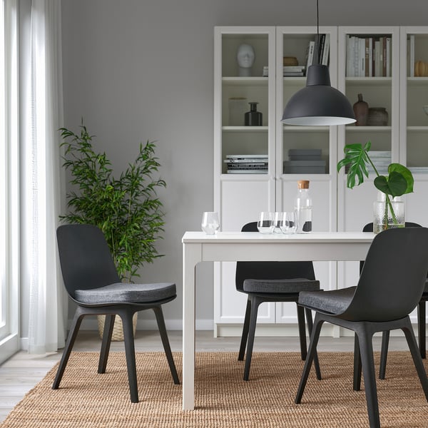 Small grey chair pads sit on black modern chairs around a white dining table, contrasting with larger green plants and open shelves behind.