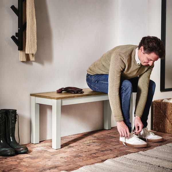A person is tying the person's trainers on a tiled floor beside a wooden bench with a white frame and a pair of black boots.